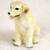 A detailed miniature figurine of a yellow Labrador Retriever puppy, designed as a decorative accent for bonsai trees. The puppy is in a sitting position, alert and facing slightly to the left with a happy expression. It features cream-colored fur with realistic textured lines, floppy ears with soft brown tips, a small black nose, and a tiny pink tongue sticking out. The paws are detailed with dark gray accents at the tips. The figurine is photographed against a soft, white textured background.