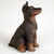 A detailed miniature figurine of a black and tan Doberman Pinscher dog with cropped ears, sitting in an alert and attentive pose. The small statue features the breed's characteristic smooth black coat with rich rust-colored markings on its muzzle, chest, and lower legs. Its ears are trimmed and stand upright, and its head is tilted slightly upward as it looks towards the right. The figurine has a subtle molded texture to simulate fur, adding a touch of realism to the tiny sculpture. This Doberman Pinscher figurine is designed as a decorative accent for bonsai trees or miniature gardens and is pictured sitting on a soft, white textured surface.