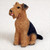 A detailed miniature figurine of an Airedale Terrier dog, designed as a decorative accessory for bonsai trees. The dog is depicted in a sitting position, looking up and to the left with its small pink tongue slightly visible. It features the breed's characteristic wiry-textured coat, with a warm tan color covering most of its body and a prominent black saddle mark on its back and tail. The figurine is realistically sculpted to show individual tufts of fur and sits against a neutral, plain off-white background.