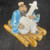 A detailed close-up of a handcrafted Chinese mud man ceramic figurine, an ornate bonsai decor piece. The figurine features an elderly man with a long, flowing dark beard, wearing a brightly glazed blue robe and white pants. He is seated on a glossy, yellowish-brown base that mimics the appearance of a bamboo raft. The man is captured in the act of playing a large, white-glazed stringed instrument, identified as a traditional Chinese banjo or pipa. His hands are delicately placed on the instrument's strings and neck. The miniature sculpture is placed on a dark, wood-grain surface, emphasizing its intricate details and traditional artistic style.