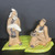 A high-resolution photograph of a two-piece Chinese ceramic figurine set, designed as a decorative accessory for a bonsai tree. The hand-crafted clay figures, in the traditional "Mudman" style, depict two men engaged in a serene, scholarly interaction. 

On the left, a standing elderly man with a long white beard and hair tied in a topknot wears tan robes with dark brown trim, holding a large white folding fan across his chest with a contemplative expression. To his right, a second man sits cross-legged on a vibrant green, leaf-textured base. He wears similar earthy-toned garments, has a cheerful, smiling expression, and holds a small white object—possibly a scroll or a tea cup—while looking up toward his companion. 

The figures are rendered in warm, matte earthy tones of tan, brown, and cream, standing out vividly against a solid dark gray background. The bright green base is divided into two interlocking pieces, textured with ridges that mimic the veins of a large leaf, providing a naturalistic foundation for these miniature traditional figures.