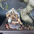 A detailed close-up shows a handcrafted ceramic Chinese figurine of an elderly man sitting and fishing, nestled at the base of a gnarled bonsai tree. The miniature fisherman, often referred to as a "mud man," features long white hair, a flowing white beard, and a calm, contemplative expression. He is dressed in a traditional tan-colored robe with dark painted accents and holds a thin wooden fishing rod with a delicate white line attached.

He is seated serenely on a dark, textured rocky outcrop, surrounded by a bed of small red, black, and green volcanic pebbles inside a deep brown ceramic bonsai pot. Directly behind him, the thick, rugged, and gnarled trunk of a mature bonsai tree rises, with a small branch of vibrant green leaves and thin aerial roots framing the scene. This traditional decorative accent adds a sense of scale, history, and peaceful storytelling to the miniature landscape of the bonsai tree.