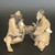 An image of a detailed, medium-sized Chinese ceramic figurine featuring two elderly wise men engaged in a strategic game of checkers or go. Handcrafted in the traditional "mud man" style, the sculpture shows the two figures seated opposite each other, connected by an arched ceramic game board that forms the center of the piece. The figure on the left, with a thick dark beard and an expressive face, holds a white game piece aloft as if contemplating a move. The figure on the right, wearing a traditional black cap and sporting a long white mustache, leans in attentively toward the board. Both figures are dressed in flowing, textured robes rendered in earthy tan and dark brown tones with a matte, unglazed finish. This traditional Chinese figurine is a classic bonsai accessory, designed to add storytelling, scale, and a sense of peaceful contemplation to a bonsai tree display or miniature tray garden.