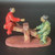 A handcrafted Chinese mud man figurine depicting a father and son playing on a seesaw, intended as a decorative accent for bonsai or penjing displays. The figure on the left is elevated high in the air, wearing a bright, glossy red glazed robe and gesturing forward with one hand. The figure on the right is at the ground level, wearing a vibrant green glazed robe and holding onto the seesaw board with both hands while looking up at his companion. Both figures feature unglazed, tan-colored clay skin and small black caps, with the figure on the right having a small white dot on his headpiece. The seesaw itself and the wide oval base are crafted from a textured, matte, reddish-brown clay. The sharp contrast between the polished, colorful glazes on the clothing and the rustic, earthy clay of the characters' skin and the seesaw highlights the traditional artisanal style of these ceramic miniatures. This scene captures a playful, serene moment, adding a sense of life and storytelling to a miniature landscape.