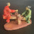 This hand-painted Chinese mud man figurine depicts a heartwarming scene of a father and son playing on a seesaw. On the left, a larger figure representing the father is clad in a vibrant, glossy red glazed robe. He is seated on one end of a rustic, tan-colored wooden-textured beam, reaching out a hand toward his son. Opposite him, a smaller figure representing the son sits on the other end of the beam, wearing a soft green glazed robe and a small black cap. 

The seesaw beam is balanced on a central support post, and the entire miniature scene is anchored to a flat, oval, terracotta-colored base. In traditional mud man style, the figures' faces and hands are left unglazed, showcasing the natural texture of the clay and expressive, hand-modeled features. This charming ceramic piece is a decorative bonsai accessory, adding a sense of narrative and tradition to a miniature garden display.