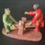 A traditional Chinese mud man figurine depicts a father and son engaging in a playful moment on a seesaw. Hand-crafted from ceramic, the figure on the left wears a glossy, glazed green robe and a small cap, leaning back as his side of the seesaw descends. The younger figure on the right is dressed in a vibrant, glazed red robe with his hair styled in traditional side buns, reaching forward with one arm extended. 

Both figures are perched on a rustic, terracotta-colored seesaw beam supported by a central post, all of which sits on a matching flat, oval-shaped base. The contrast between the matte, unglazed finish of the skin and seesaw and the high-shine glaze of the colorful robes highlights the intricate detail of this decorative bonsai accessory. This small piece is designed to bring a sense of life, story, and tradition to a bonsai tray landscape or an indoor garden display.