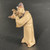 A detailed side-profile photograph of a handcrafted Chinese ceramic figurine, model F-035, often used as a decorative "Mudman" for bonsai displays. The figurine depicts a man in a standing, contemplative pose, leaning slightly forward. He is dressed in traditional, flowing robes of a light tan or sandy beige color, featuring subtle dark brown glazed accents that highlight the deep folds and drapes of the fabric. His wide sleeves are gathered as he holds his hands near his chest in a deliberate, scholarly gesture. On his head, he wears a dark brown peaked cap adorned with a small, white circular bead on the front. The figurine's face has simple, hand-sculpted features that convey a focused and serene expression. The piece has a rustic, earthy texture with a mix of matte surfaces on the robes and a slight sheen on the glazed hat. Set against a dark, minimalist background, this "man standing" figurine is designed to add a sense of scale, narrative, and cultural heritage to a miniature landscape or indoor decor.