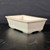 An angled side view of a 7-inch glazed cream rectangle Yixing ceramic bonsai pot (model No. 2514b) showcased on a dark, textured surface. The pot features an off-white or light beige glossy finish with a subtle crackle glaze pattern throughout. Its design is classic and functional, with sides that slope slightly outward from the base towards a prominent, rounded rim. A decorative horizontal ridge or "belt" wraps around the lower portion of the pot just above its four small, integrated rectangular feet that provide drainage clearance. The ceramic pot is set against a dark charcoal-grey background, which highlights its smooth curves and soft cream color, while the dark, wood-textured surface below captures a faint reflection of the vessel.
