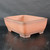 A high-angle studio photograph of a 14-inch unglazed Yixing bonsai pot, model YX1094. The rectangular vessel is crafted from warm terracotta clay, featuring a smooth matte surface with subtle, dark-brown shading along its rim and base. Its sides are slightly bowed, leading to rounded corners and a flat top edge. The pot is elevated by four short, sturdy rectangular feet at the corners. Set against a dark, minimalist background on a slightly reflective black surface, the soft lighting emphasizes the earthen textures and classic design of the container, which is ideal for various bonsai tree styles.