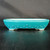 A shallow, 12-inch oval Yixing ceramic bonsai pot sits atop a dark, wood-grain surface against a neutral, dark gray background. The pot is coated in a vibrant teal-turquoise glaze with a glossy, fine crackle finish that reflects light. Its most distinctive feature is the series of raised, circular "rivets" or bumps arranged in staggered rows along the lower half of its exterior, giving it a textured, traditional appearance. The pot has a white-lined interior visible at its smooth, rounded rim. It is supported by four small, decoratively carved feet that add a touch of elegance to its sturdy, low-profile design.