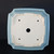 A detailed overhead view shows the bottom of a square Yixing ceramic bonsai pot, highlighting its functional and decorative features. The pot is finished in a smooth, light blue glaze that coats the exterior and its tiered, mokko-style rim with indented corners. The flat, unglazed base is an off-white ceramic color and features five precisely cut openings: a large central drainage hole for water management and four smaller wire holes, one in each quadrant, used for securing a bonsai tree with training wire. Near the top of the unglazed area, an impressed square potter's seal indicates the maker’s mark. The pot's clean lines and soft blue hue contrast sharply against the dark black background.