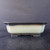 A detailed studio photograph of a 9-inch square Yixing ceramic bonsai pot. The pot features an elegant two-tone gradient glaze, with a dark charcoal or deep brown flared rim that smoothly transitions into a pale, sage green or celadon color across the main body. 

The square design is softened by beveled corners and slightly tapered sides, giving it a refined and traditional look. The pot is elevated on four integrated, low-profile feet, creating a subtle arch underneath its base. Set against a minimalist dark gray background and resting on a black, wood-textured surface, the lighting highlights the smooth finish and subtle color shift of the ceramic. This glazed Yixing pot is a classic choice for showcasing a variety of bonsai tree species, blending functional design with aesthetic appeal.