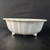 A side-profile photograph shows a 17-inch white glazed Yixing bonsai pot. The large, shallow pot features a wide, slightly oval shape with an elegant scalloped or wavy rim. Its exterior walls are decorated with soft, vertical fluting that adds a classic texture to the design. The pot is elevated on four small, decorative, scrolled feet that provide both stability and aesthetic appeal. The glossy white ceramic glaze gives the piece a clean, sophisticated finish that catches the light subtly. It is positioned on a dark, textured surface against a minimalist black background, highlighting the pot's bright color and smooth, handcrafted details.