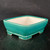 A long, rectangular Yixing ceramic bonsai pot with a vibrant, glossy teal-green glaze on its exterior. The pot's design features slightly tapered walls and softly rounded corners, leading down to a decorative flared base. It sits elevated on four thick, blocky feet, one at each corner, which are also finished with the same rich green glaze. The interior of the pot is left unglazed, showing a smooth, clean, off-white ceramic surface. The pot is captured at a three-quarter angle on a dark, textured surface against a solid black background, which makes the bright green color pop and highlights the smooth, reflective quality of its glaze. This 7-inch pot is designed for displaying a small-to-medium-sized bonsai tree.
