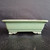 A side view of a 7-inch rectangular glazed Yixing bonsai pot in a soft celadon green. The pot features a prominent, slightly overhanging top rim and a body that gently tapers toward the base. It stands on four decorative, cloud-style scroll feet that have a slightly weathered appearance with touches of exposed clay. The smooth, light green glaze is uniform with subtle, fine dark speckling characteristic of high-quality ceramic. The pot sits on a dark, wood-grain surface against a plain, dark gray fabric backdrop, emphasizing its traditional aesthetic and elegant proportions.
