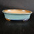 A 9-inch glazed oval Yixing bonsai pot is featured in this studio photograph, showcasing its elegant design and serene color palette. The exterior of the shallow pot is finished in a vibrant turquoise-blue glaze with a subtle, mottled texture. A contrasting darker blue glazed band runs along the bottom edge, just above the small, unglazed tan feet that provide essential drainage and elevation.

The pot has a classic oval shape with a gently flared rim that reveals a natural, unglazed tan ceramic interior, providing a warm, earthy contrast to the blue exterior. It is presented on a dark, textured wooden surface against a solid black background, which makes the pot’s colors and smooth finish pop. This high-quality Yixing ceramic pot is designed to complement a wide variety of bonsai tree species, blending traditional craftsmanship with a modern aesthetic.
