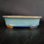 A professional product shot of a 9-inch glazed oval Yixing bonsai pot (No. 2329A). The pot has a sophisticated oval shape with walls that flare slightly outwards from a recessed base towards a thin, distinct rim. It is finished in a light teal or grayish-blue glaze that features a delicate, fine crackle pattern across its surface.

A darker, deeper cobalt blue glaze accents the bottom edge of the pot, creating a beautiful gradient effect. The top edge of the rim and the four small, blocky feet at the base are left unglazed, showing the natural, warm, earthy-brown color of the Yixing clay. The pot is positioned on a dark, subtly textured black surface, with its glossy finish reflecting the light against a solid black backdrop. This classic, handcrafted ceramic vessel is designed to complement various bonsai styles with its elegant form and rich color palette.