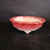 A close-up photograph displays a 9-inch glazed round red Yixing bonsai pot, item number 1092. The pot features a decorative scalloped or lobed rim and small, ornate feet that elevate it slightly. Its most striking feature is a vibrant gradient glaze that transitions from a deep, glossy cherry red at the top rim to a soft pink and creamy off-white toward the base. The interior of the bowl is unglazed or a light beige color, providing a neutral backdrop for a future plant. The pot is positioned on a dark, textured wooden surface against a solid black background, making the rich red color and elegant curves of the ceramic piece stand out.
