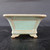 A side-profile view of a 5-inch green glazed square Yixing bonsai pot, model number 1841a. The pot is finished in a serene, pale seafoam green glaze with a subtle speckled texture and warm, brownish-tan accents along the rim, edges, and recessed rectangular side panel. Its design features a wide, flared top rim that tapers gracefully down to a footed base with elegant, curved feet. The pot is positioned on a dark, textured surface against a plain black backdrop, highlighting its architectural shape and smooth ceramic finish, making it a classic choice for showcasing a small bonsai tree.