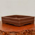 An eye-level, three-quarter view of a 9-inch rectangular Yixing bonsai pot with a classic unglazed finish. The pot is crafted from deep, earthy reddish-brown clay, displaying a smooth, matte texture characteristic of authentic Yixing pottery. 

A decorative double-line horizontal band wraps around the middle of the pot's exterior, adding a subtle touch of detail to its minimalist design. The pot is shallow with softly rounded corners and features small, integrated feet at the base to provide stability and essential drainage for a bonsai tree. 

The pot sits on a highly polished, reddish-brown wooden surface with a distinctive, gnarled live edge in the foreground. The background is a plain, light-colored wall, which helps to highlight the clean lines and rich color of the unglazed ceramic.