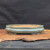 A shallow, oval-shaped Yixing ceramic bonsai pot sits centered on a rustic wood slab. The pot features a smooth, glossy glaze in a muted grayish-green or celadon color, with subtle variations and highlights reflecting from its curved surface. Its design includes a slightly rounded body, a thin defined rim, and four small, integrated feet that lift it slightly off the surface. The interior of the pot is unglazed, revealing the natural, warm tan-orange color of the clay. The planter is displayed on a thick, dark-grained wooden base with a natural live edge, which rests on a dark foreground against a solid black fabric backdrop, emphasizing the bonsai pot's elegant form and earthy color palette.
