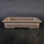 A studio photograph of an unglazed Yixing bonsai pot, approximately 10.5 inches long, showcasing its classic rectangular design. The pot is crafted from a smooth, matte brownish-grey clay with a wide, level rim. A decorative horizontal recessed panel wraps around the exterior, adding subtle architectural detail. It features traditional cut-out feet on the bottom to provide drainage and airflow. The pot is shown empty and centered on a dark, textured wooden surface against a plain black background, emphasizing its clean lines and earthy aesthetic.