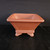A 7-inch unglazed square cascade Yixing bonsai pot crafted from reddish-brown terracotta clay. The pot features a wide, flat rim and sides that taper downward toward a smaller base, creating the deep vessel characteristic of a cascade-style pot. Each of the four sides is decorated with a recessed panel and beveled edges, adding geometric depth. Near the bottom, two horizontal raised bands wrap around the base, which sits on four decorative, outward-curving feet. The pot is centered on a dark, textured black surface against a solid dark background, with soft lighting highlighting its matte finish and architectural details.