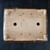 A top-down view of the underside of a 12-inch shallow rectangular Yixing ceramic bonsai pot. The unglazed clay base has a light tan color, while the outer rim is finished with a glossy dark brown glaze. The pot features four L-shaped feet at each corner for stability. Functional details include two large circular drainage holes on the left and right, and four smaller wire holes positioned in a rectangular pattern between them. In the center, there are three impressed maker’s marks or chops: a square stamp with Chinese characters, an oval-shaped seal, and a stylized mountain-like logo above them. The pot rests on a dark, textured black surface, showcasing the authentic texture and details of the ceramic craftsmanship.
