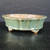 A 7-inch glazed Yixing bonsai pot, model YX1030-1C, featuring an elegant, pale seafoam green glaze. The pot has a distinctive oval shape with a scalloped, lobed exterior that creates a series of soft vertical ridges and a gently flared rim. Its glossy finish reflects light, and the glaze subtly thins at the edges, allowing the warm, reddish-brown tones of the natural Yixing clay to show through. 

The pot is elevated on four small, integrated feet that raise it slightly off the surface. While the exterior is beautifully glazed, the interior remains unglazed, revealing the raw, porous terracotta-colored clay—a hallmark of high-quality bonsai pots that promotes healthy root growth through better aeration. The container is photographed against a dark, minimalist background on a wood-grained surface, highlighting its delicate color and handcrafted details.