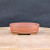 A detailed studio photograph of a 4.5-inch unglazed oval Yixing bonsai pot. The pot is made from a traditional reddish-brown clay with a smooth, matte finish that is characteristic of high-quality Yixing pottery. It features a classic oval shape with a flat, clean rim and a subtle raised horizontal band encircling the lower third of the body. The pot sits on four low, integrated feet that provide a slight lift from the surface. It is centered on a light-colored, rustic wooden block or turntable, set against a dark, textured charcoal background that highlights the warm, earthy tones of the clay. This professional-grade bonsai pot is designed for small trees or succulents, offering the porous breathability essential for healthy root development.