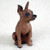 A small, detailed figurine of a red-brown Miniature Pinscher dog, designed as a decorative accessory for a bonsai tree or terrarium. The figurine is captured in a sitting position, looking alertly toward the upper right with its large, pointed ears standing upright. Its dark, glossy eyes and small black nose give it an expressive and lifelike face. The surface of the reddish-brown resin is textured to mimic the short fur of a Min Pin. The dog is posed on a soft, off-white surface that provides a neutral background for the miniature sculpture.