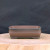 A rectangular Yixing clay bonsai planter with a rustic, hand-finished aesthetic is centered on a light-colored, polished wood slab. The unglazed ceramic pot features a deep chocolate brown color with dark, charcoal-like accents along the thick, rounded upper rim and the four low, broad feet. Two subtle horizontal bands wrap around the middle of the pot's exterior, adding a simple decorative touch. The planter’s interior is a uniform matte brown, indicating its porous nature suitable for plant health. The entire piece is set against a solid, textured black fabric background, which provides a high-contrast backdrop that emphasizes the earthy tones and traditional design of the bonsai pot.