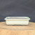 A horizontal, close-up shot features a seven-inch rectangular bonsai pot with a smooth, cream-colored glaze. The planter has a slightly flared rim and a decorative, recessed rectangular panel on its front face, adding a touch of classic detail. It sits on four short, integrated feet that reveal a dark, unglazed ceramic base, which matches the dark brownish-gray interior of the pot. 

The planter is centered on a thick, light-toned wooden cross-section slab, showcasing the natural grain and texture of the wood. A solid, dark charcoal-grey background provides a stark, minimalist contrast, emphasizing the clean lines and soft color of the ceramic pot. This elegant cream-glazed planter is designed specifically for small bonsai trees or other miniature indoor plants.