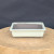 A seven-inch rectangular cream glazed bonsai pot sits centered on a rustic, circular wooden log display stand against a solid dark gray backdrop. The ceramic planter has a smooth, pale cream-colored glaze covering its exterior, featuring a subtle raised rim and a decorative recessed panel on the side. The interior and the four small feet at the base are unglazed, showing a contrasting dark brown clay body. This high-quality ceramic planter is designed for small bonsai trees, offering a clean and elegant aesthetic for indoor or outdoor display.