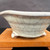 A side-view photograph shows a 5.5-inch round Tokoname bonsai pot, expertly crafted by Kakuzan (Kakuyuki Watanabe). The ceramic pot features a beautiful soft gray crackle glaze, with a network of fine, dark lines across its surface creating an aged, artisanal aesthetic. Its design includes a wide, flared rim and it is supported by three small, rounded feet that show hints of the underlying reddish-brown clay at their base. The pot is positioned on a light-colored, polished wooden display stand, set against a dark, minimalist background that emphasizes the delicate texture and elegant proportions of the vessel.