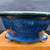 A side-view photo of a handcrafted 5.25-inch round Tokoname bonsai pot by renowned Japanese artist Kakuzan (Kakuyuki Watanabe). The pot features a vibrant deep blue glaze with a unique crystalline texture that creates a shimmering, starry effect across its surface. A slightly darker, midnight-blue band highlights the subtly flared upper rim. The pot is supported by three small, rounded feet that transition from the blue glaze to natural, unglazed pale clay at the base. It is displayed on a polished light-wood block against a dark, out-of-focus background, emphasizing the intricate details and rich color of this traditional ceramic piece.