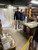 A wide-angle indoor shot shows two men standing together inside a professional bonsai pottery workshop in Tokoname, Japan. On the right is Kouji Watanabe, a master Japanese bonsai potter, wearing a dark blue quilted vest over a long-sleeved shirt and beige trousers. He is smiling warmly next to a visitor, a man in a black baseball cap, black hoodie, and grey jeans, who has his arm around Watanabe's shoulder in a friendly gesture. 

The studio environment is filled with the tools and products of the bonsai trade. To the right, tall wooden shelves are stocked with various ceramic bonsai pots in different glazes and shapes. A stack of blue plastic crates sits against a wall behind them, and a large wooden worktable in the center is covered with a clear protective sheet over maps or design documents. In the foreground, a stack of brochures titled "BONSAI POTS" sits on a low wooden bench. The background reveals a workspace divided by translucent plastic sheeting, typical of a pottery studio. This setting is where Kouji Watanabe crafts his renowned pieces, such as his unglazed red clay tall pots featuring intricate dragon reliefs and traditional oni feet, embodying the rich heritage of Tokoname bonsai pottery.
