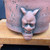 A detailed close-up of an unglazed red clay Tokoname tall bonsai pot, meticulously crafted by artist Kouji Watanabe. The image highlights a prominent relief carving of a fierce "Oni" (Japanese demon) face that serves as one of the pot's feet. This intricately sculpted Oni mask features two pointed horns, a furrowed brow over bulging eyes, a broad nose, and a grimacing mouth revealing small teeth. The chin of the mask rests firmly on a light-colored wooden display stand. The body of the pot exhibits a rich, earthy reddish-brown hue characteristic of traditional Tokoname pottery, with a rustic and aged texture defined by numerous small pits and dark, irregular indentations. The dark, neutral background provides a sharp contrast, drawing focus to the detailed craftsmanship and weathered appearance of the unglazed clay.