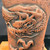 A detailed close-up of a handcrafted Tokoname bonsai pot by Kouji Watanabe, made from unglazed red clay. The pot features a dynamic and highly detailed dragon relief carving that wraps around its tall, cylindrical body. The dragon's head is shown in profile with fierce features, sharp teeth, and a piercing eye, with its long, textured mane flowing backward. Its body is covered in intricately carved, overlapping scales, and a powerful claw with sharp talons is prominent below the head. The unglazed clay has a rustic, porous texture with fine pits and a rich earthy tone, emphasizing the artisanal quality of this traditional Japanese pottery piece.