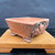 A 4.5-inch rectangular Tokoname bonsai pot crafted from unglazed red clay by artist Kouji Watanabe. The pot features a prominent, hand-carved relief of a dragon on its side, adding a dynamic and traditional artistic element. Its body has a rustic, textured finish with dark, pitted speckles throughout, giving it an aged, natural look. The pot is supported by four unique, sculpted feet that resemble animal claws. It is displayed on a light-colored wooden stand against a dark, mottled background, highlighting the rich reddish-brown hue of the clay and the intricate details of the dragon sculpture.
