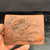 An unglazed red clay plaque, approximately 4.25 inches wide, featuring an intricate hand-carved relief of a traditional East Asian dragon. The dragon is masterfully rendered with fine details, including individual scales, flowing whiskers, sharp horns, and taloned claws as it winds through stylized, swirling clouds. The plaque is made of authentic Tokoname clay, showing a warm terracotta hue with darker shadows in the deep recesses of the carving to emphasize depth. In the top-left corner, Japanese kanji characters and a rectangular artist’s seal mark indicate the piece was crafted by Kouji Watanabe. The plaque is held between a person's fingers against a dark, textured background, highlighting its artisanal quality and compact size, suitable for use as a bonsai display accessory.