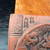 A high-detail close-up of a corner of a square Tokoname unglazed red clay bonsai plaque. On the left is a prominent rectangular artist's seal containing the characters "幸峰" (Kouhou), the art name of artist Kouji Watanabe. To its right are smaller vertical inscriptions referencing the "Kakuzan" kiln. The right side of the frame showcases a portion of a finely hand-carved relief depicting a dragon with intricate scales and textured skin. The earthy terracotta clay has a matte, granular finish, and the plaque rests on a dark, wood-grained surface next to a light-colored wooden block.