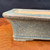 A detailed close-up shot of an 8.5-inch Reiho (Katsushi Kataoka) Blue Speckled Rectangle Bonsai Pot from the Seizan Kiln. The ceramic pot features a sophisticated, textured glaze with dense blue and dark-brown speckles over a soft, seafoam-green base. The rectangular shape is defined by a thick, rounded rim with a brownish-bronze accent along its upper edge. The interior is unglazed, revealing a light tan clay. The pot is showcased on a polished, reddish-brown wooden display table against a solid black background, emphasizing the intricate details and artisanal quality of the Seizan Kiln's craftsmanship.