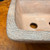 A detailed, high-angle close-up showcases the corner of a 6-inch Reiho Katsushi Kataoka bonsai pot from the Seizan Kiln. The unglazed ceramic pot is notable for its thick, heavily textured rim, which displays a rugged, pebbled surface reminiscent of weathered rock or tree bark. Its color is a complex blend of earthy gray and warm brown tones. The smoother interior of the pot shows a circular drainage hole and an authentic potter's mark hand-written in white Japanese characters. The pot is positioned on a polished, natural wood surface, emphasizing its handcrafted, artisanal quality.