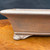 A detailed side-profile view shows a 9.75-inch unglazed rectangular bonsai pot, handcrafted by artist Katsushi Kataoka at the renowned Seizan Kiln. The pot is made from earthy, reddish-brown clay with a matte, finely textured surface that displays subtle mottling and speckles. 

Its design features a thick, elegantly flared rim and a horizontal decorative ridge near the base. A key highlight is the ornate "cloud foot" design, where the corner foot is sculpted with several smooth, rounded lobes, adding a traditional Japanese aesthetic to the piece. The pot rests on a polished, orange-brown wooden surface with a rich grain, set against a dark, solid background that emphasizes the pot's natural color and artisanal detail.
