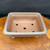 A detailed view of a 9.75-inch Reiho Katsushi Kataoka deep unglazed rectangle bonsai pot from the Seizan Kiln. This handmade ceramic pot features a classic rectangular shape with notched corners and a flat rim. The clay has a warm, reddish-brown hue with a matte, slightly weathered texture on the exterior. Inside, the bottom of the pot shows five drainage holes and a white handwritten inscription that reads '8号'. The pot is resting on a polished natural wood slab with an intricate grain, set against a dark, neutral background.