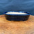 An eye-level, slightly angled shot showcases a 10.25-inch dark blue oval bonsai pot, a product of the Seizan Kiln by artist Katsushi Kataoka (Reiho). The pot is finished in a deep, glossy navy blue glaze that catches the light, highlighting its smooth surface and two subtle horizontal stepped ridges that encircle the outer wall. It features a gently flared rim and sits on small, integrated feet that provide a slight lift. The interior is unglazed, revealing a light tan ceramic texture. The bonsai pot is centered on a polished wooden slab with a rich, warm-toned burl grain. A solid, dark blue fabric serves as a simple, non-distracting background that makes the pot's color pop.