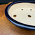 A close-up, high-angle photograph showcasing a 10.25-inch dark blue oval bonsai pot crafted by artist Reiho Katsushi Kataoka from the Seizan Kiln. The pot features a deep, lustrous cobalt blue glaze on its smooth outer rim and exterior wall, which reflects a bright light source. The interior of the ceramic pot is unglazed, revealing a fine-textured, light tan clay surface. Four circular drainage holes are visible at the bottom of the pot, along with some white chalk markings. The entire piece rests on a polished, warm-toned burl wood surface with a rich, swirling grain. This artisanal container is designed for both the aesthetic display and the healthy growth of a bonsai tree.