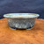 A detailed view of a 6-inch Reiho (Katsushi Kataoka) mottled blue-green oval bonsai pot from the Seizan Kiln. This ceramic bonsai container is characterized by its elegant oval shape, flared rim, and a rich, speckled glaze that combines deep indigo with flecks of pale seafoam green. The pot sits on small, integrated feet, with the unglazed tan clay visible at the very bottom edge and throughout the interior. It is displayed on a rustic, polished wood surface with a prominent swirling grain, set against a dark, minimalist fabric background that highlights the intricate textures and colors of the glaze. This high-quality Japanese bonsai pot exemplifies the traditional artistry and craftsmanship of the Seizan Kiln.