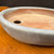 An extreme close-up shot of a 5.75-inch vintage Seizan Kiln glazed oval bonsai pot, showcasing its unique texture and color. The pot's exterior is coated in a muted, mottled blue-grey glaze with soft lavender undertones and subtle dark speckles, giving it a weathered, previously-used character. The wide, rounded rim leads into an unglazed interior, revealing the warm, reddish-brown terracotta clay. A circular drainage hole is visible on the flat bottom of the pot. The vessel is placed on a rustic, warm-toned wooden surface with a fine grain, while a dark, out-of-focus background makes the light-colored ceramic stand out.